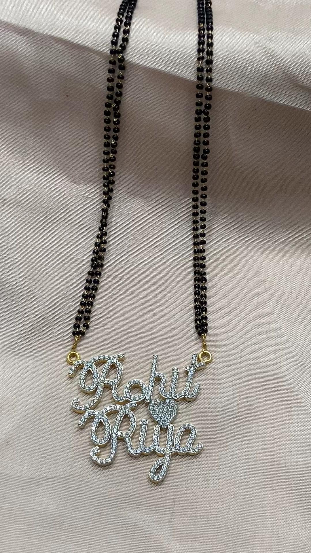 his Mangal Sutra is more than just a piece of ornament — it’s a declaration of forever. The dual name design, delicately linked with a heart-shaped motif, radiates warmth and intimacy. Adorned with round-cut American Diamonds that twinkle like shared dreams, this mangalsutra reflects the sparkle of love in its purest form. The double-layered black beaded chain adds a traditional touch, symbolizing protection, trust, and commitment, while blending effortlessly with modern aesthetics.